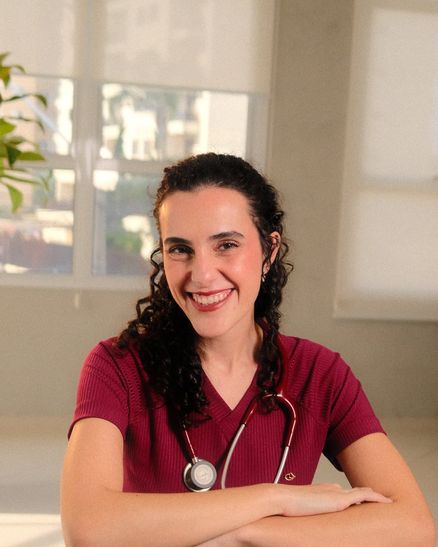 A Dra. Anna Luiza Pollon sorrindo, de jaleco vinho e estetoscópio, apoiada numa mesa junto à janela.